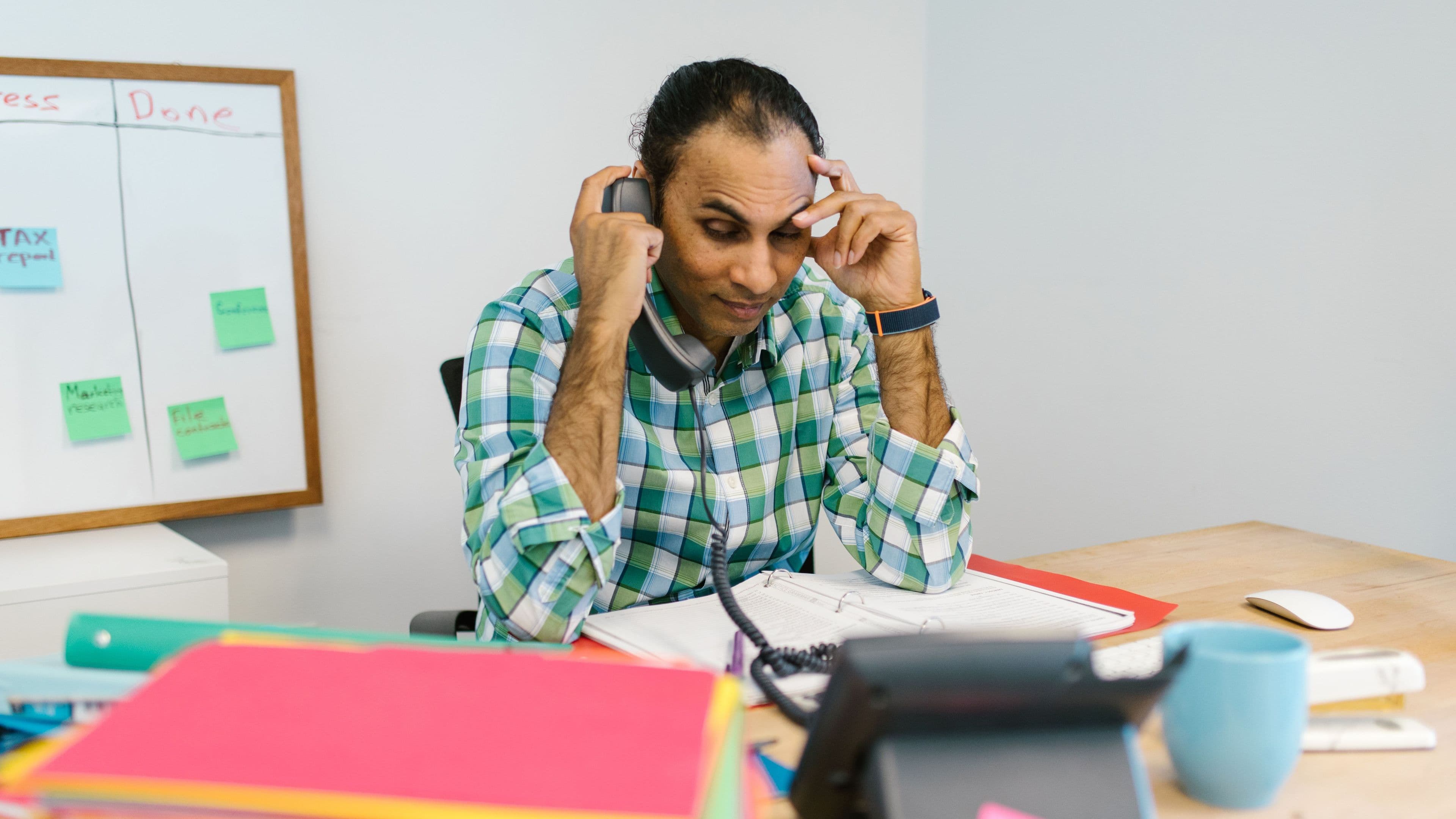 A professional holding his head at his desk in front of a task board