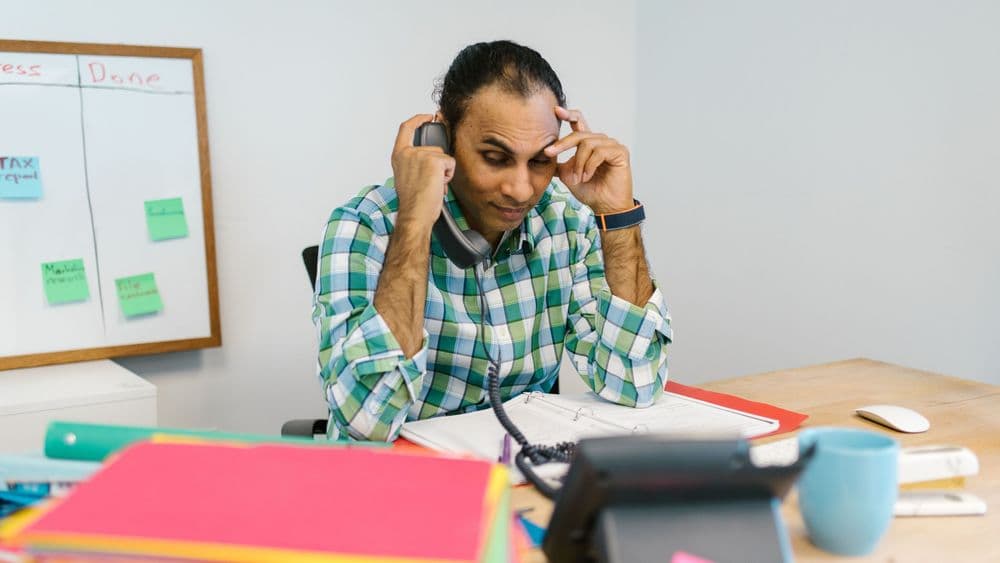 A professional holding his head at his desk in front of a task board