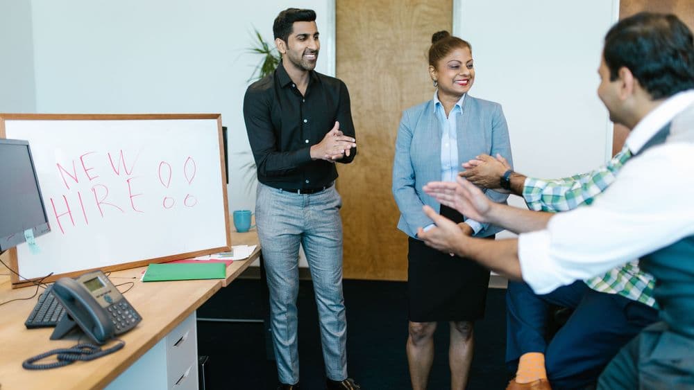 Colleagues welcoming a new hire beside a whiteboard reading New Hire