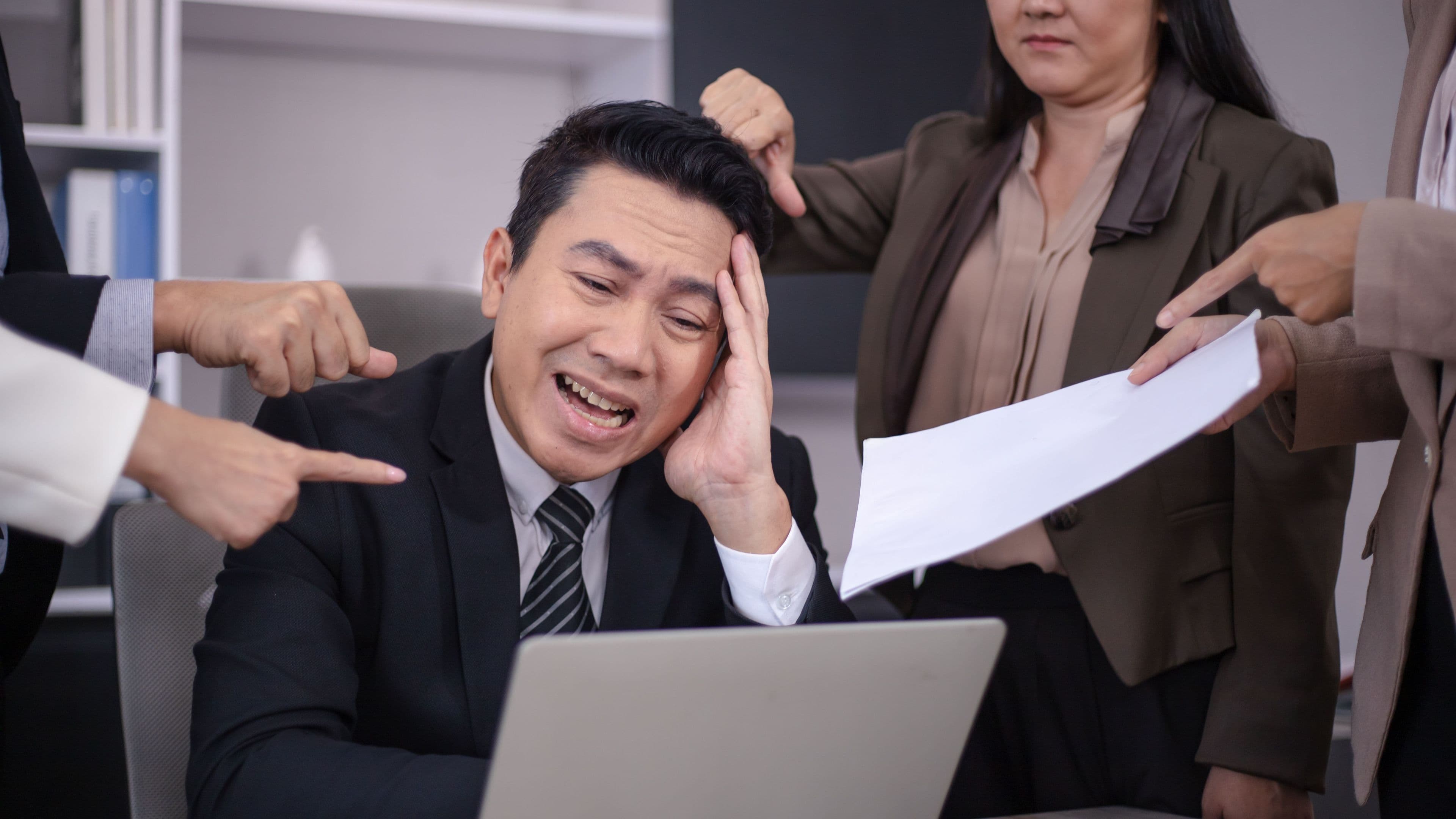 A stressed employee at a laptop as a colleague presses him with paperwork